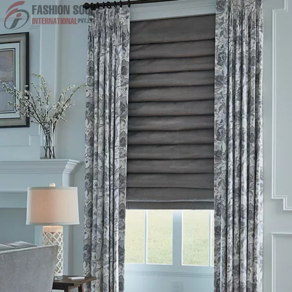 A window with a grey Roman shade partially pulled down, framed by floral-patterned curtains in shades of grey and white. To the left, there is a table lamp with a white lampshade, a side table, and a decorative branch arrangement on a mantelpiece in a cozy, well-decorated room.