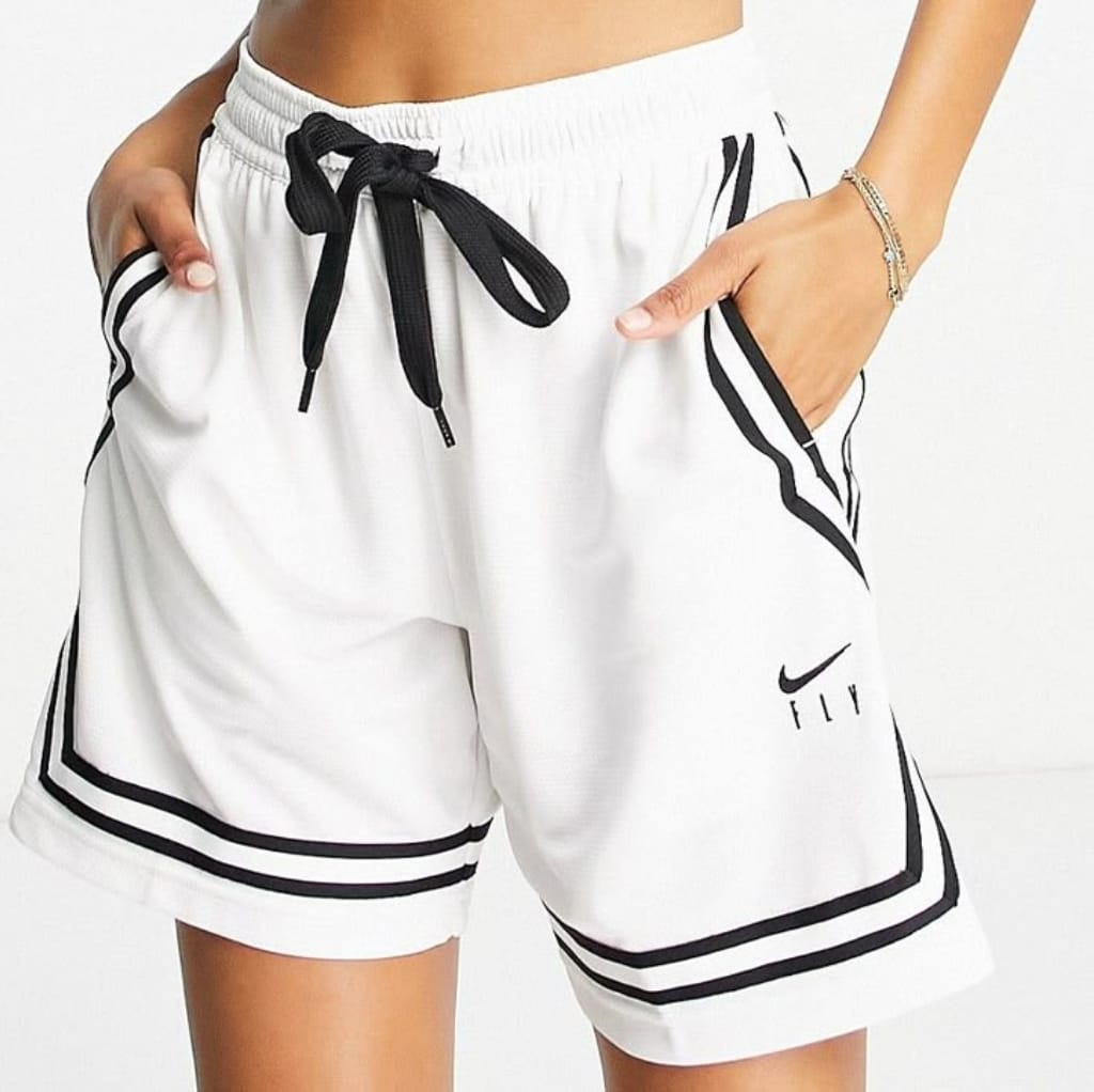 White Nike shorts with black trim and a black drawstring, featuring the Nike logo on the left leg and two black stripes along the bottom hem. The model has one hand in the pocket and is wearing bracelets on her wrist. Best Nike Sports Wear