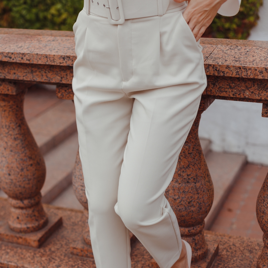 Close-up image of a person wearing elegant, high-waisted off-white pants with pleats, standing outdoors next to a rust-colored stone railing.
