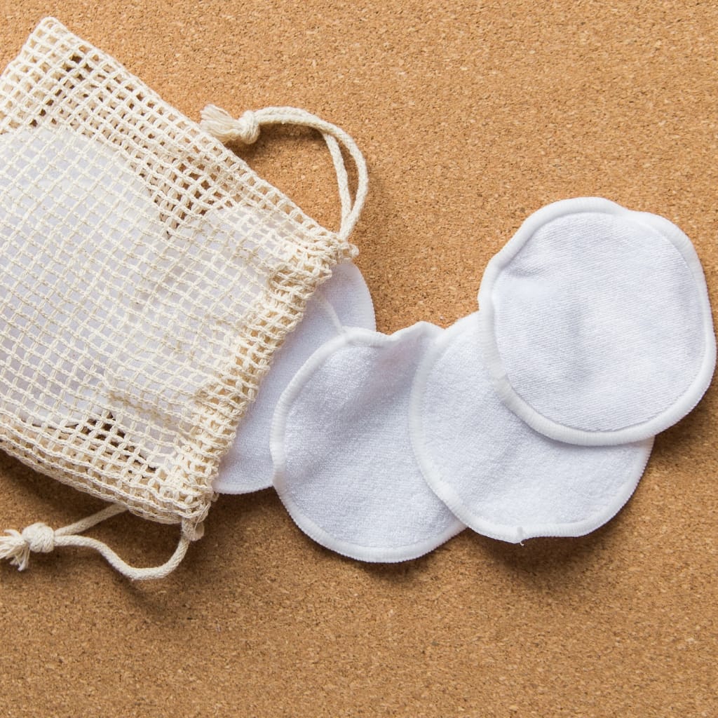 A string of white facial cotton pads next to a reusable mesh bag, on a cork surface.