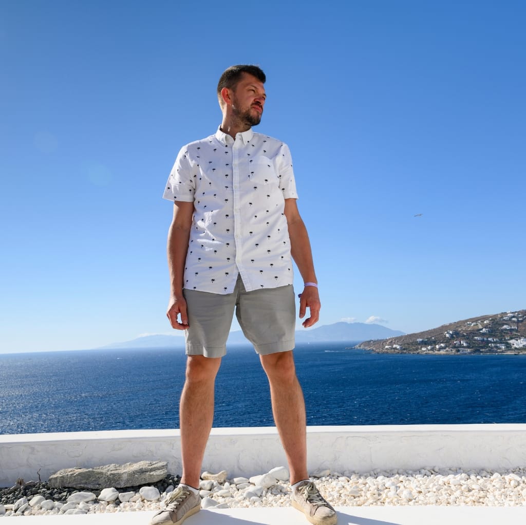 A man standing outdoors by the water, wearing a white short-sleeve shirt with small dark patterns, light gray shorts, and beige sneakers, under a clear blue sky.