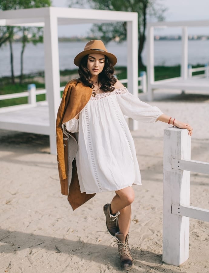 A woman wearing a white cotton babydoll dress, a brown hat, and rugged boots, standing outdoors near a white fence with water and trees in the background.