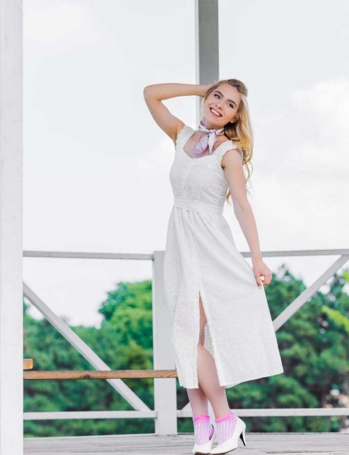A young woman standing outdoors on a balcony, wearing a white sleeveless dress with a slit, pink socks, and white high heels. She has blonde hair and is smiling, with one hand behind her head.