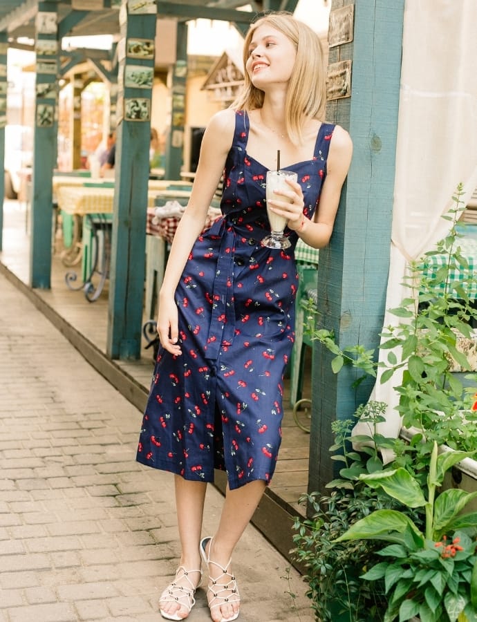 A young woman in a navy blue pinafore dress with a cherry print, standing outdoors near a wooden structure, holding a milkshake with a straw, wearing white sandals.