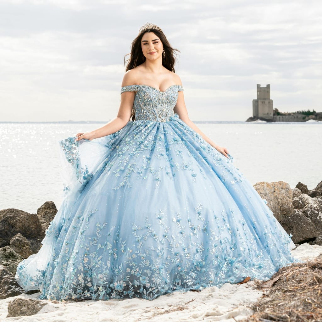 A young woman in a stunning blue off-shoulder ball gown with intricate floral embroidery, standing on a rocky beach with a castle in the background.