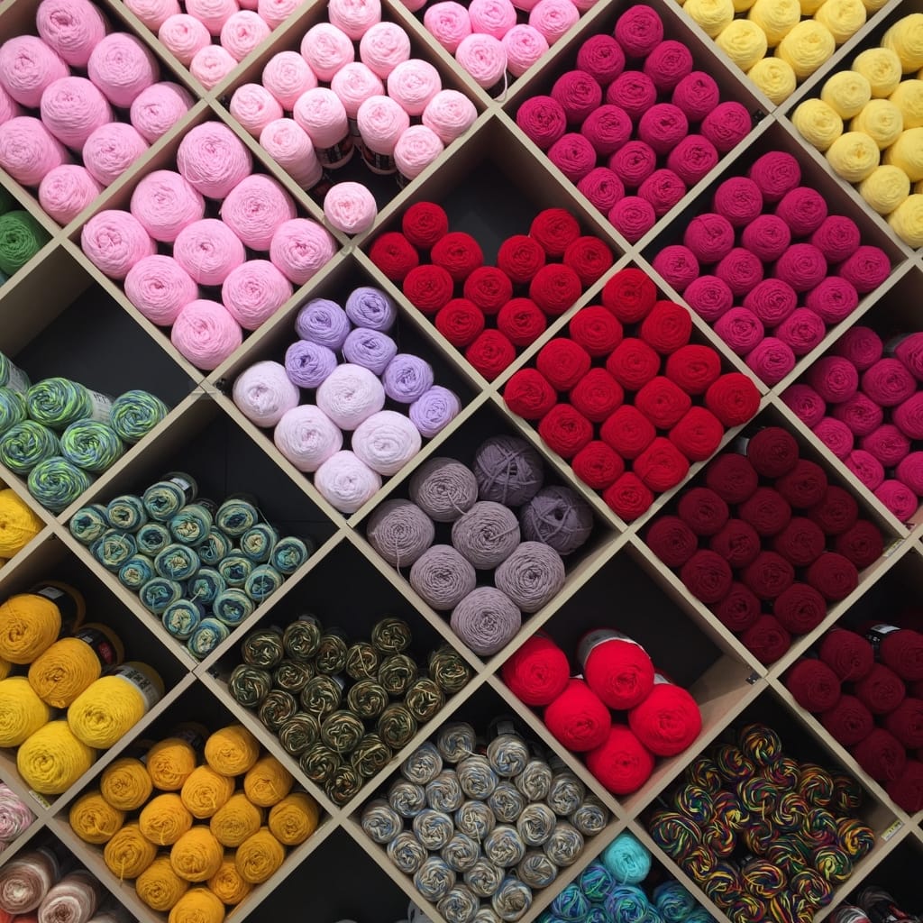 A neatly organized display of colorful yarn balls in cubby-style compartments, featuring shades of pink, purple, yellow, red, green, and multicolored varieties