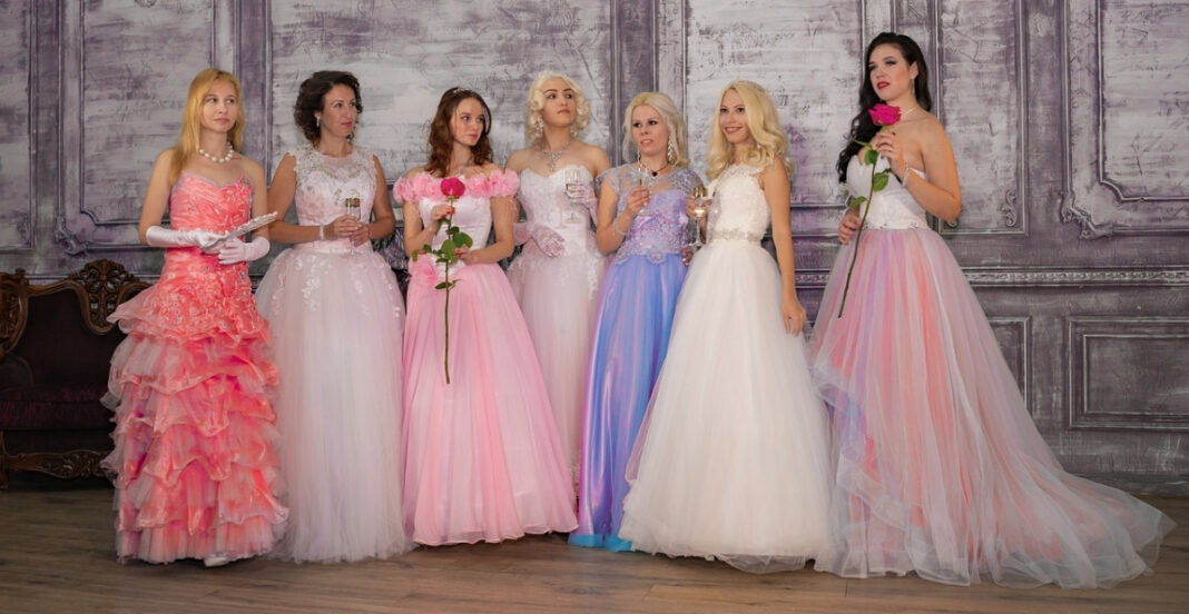 A group of seven women in elegant prom dresses standing indoors against a grey ornate wall, holding roses and glasses, with some smiling and looking away.