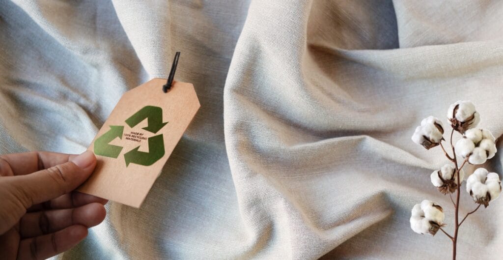 A close-up of a hand holding a beige clothing tag with a green recycling symbol and the text "MADE BY RECYCLE.ME" on a piece of light-colored fabric, with a small branch of cotton flowers resting on the fabric.