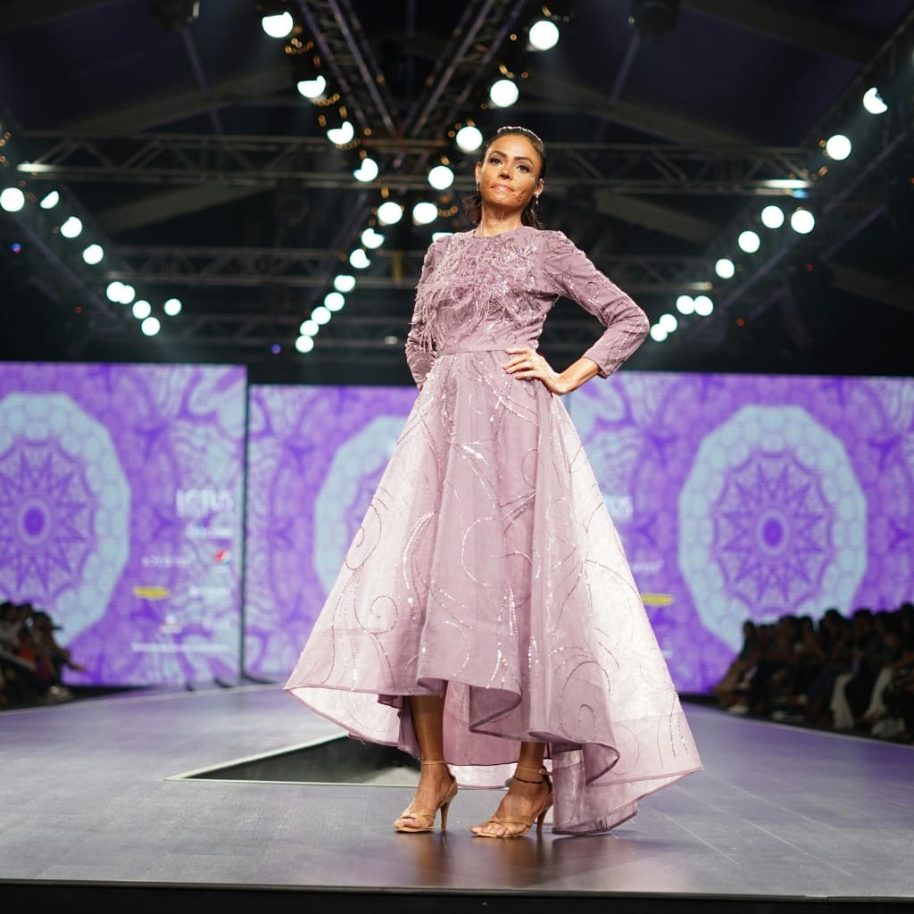 A model walking down a runway in a lavender organza silk dress with intricate embroidery, under bright stage lights, with an audience watching on.