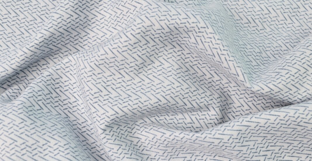 Close-up image of light-colored Sea Island cotton fabric with fine, small, dark dashed lines forming a subtle, intricate pattern.