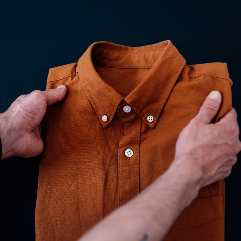 A person folding a rust-colored short sleeve button-down shirt against a dark background.