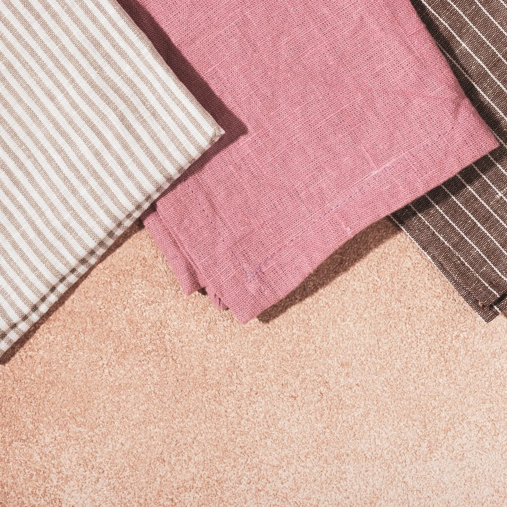 Three folded fabrics with different patterns and colors, placed on a sandy surface — one with beige and white stripes, one solid pink, and one dark brown with white stripes.
