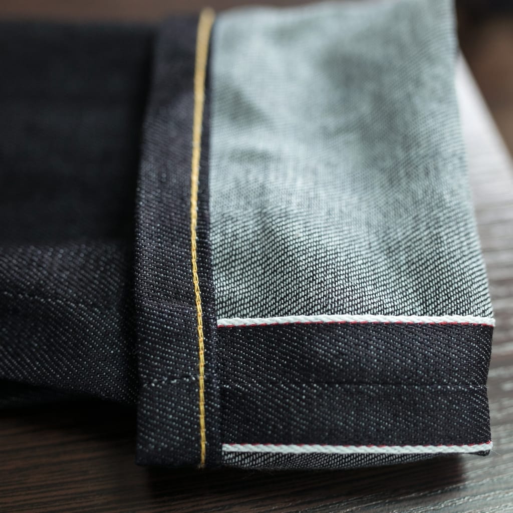 Close-up of the edge of a pair of selvedge denim jeans, showing the tightly woven selvage edge with white and red stitching, and a yellow line running along the selvage.