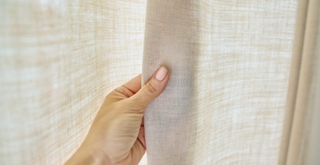 A hand gently holding back a light-colored fabric curtain, revealing more of the same material.
