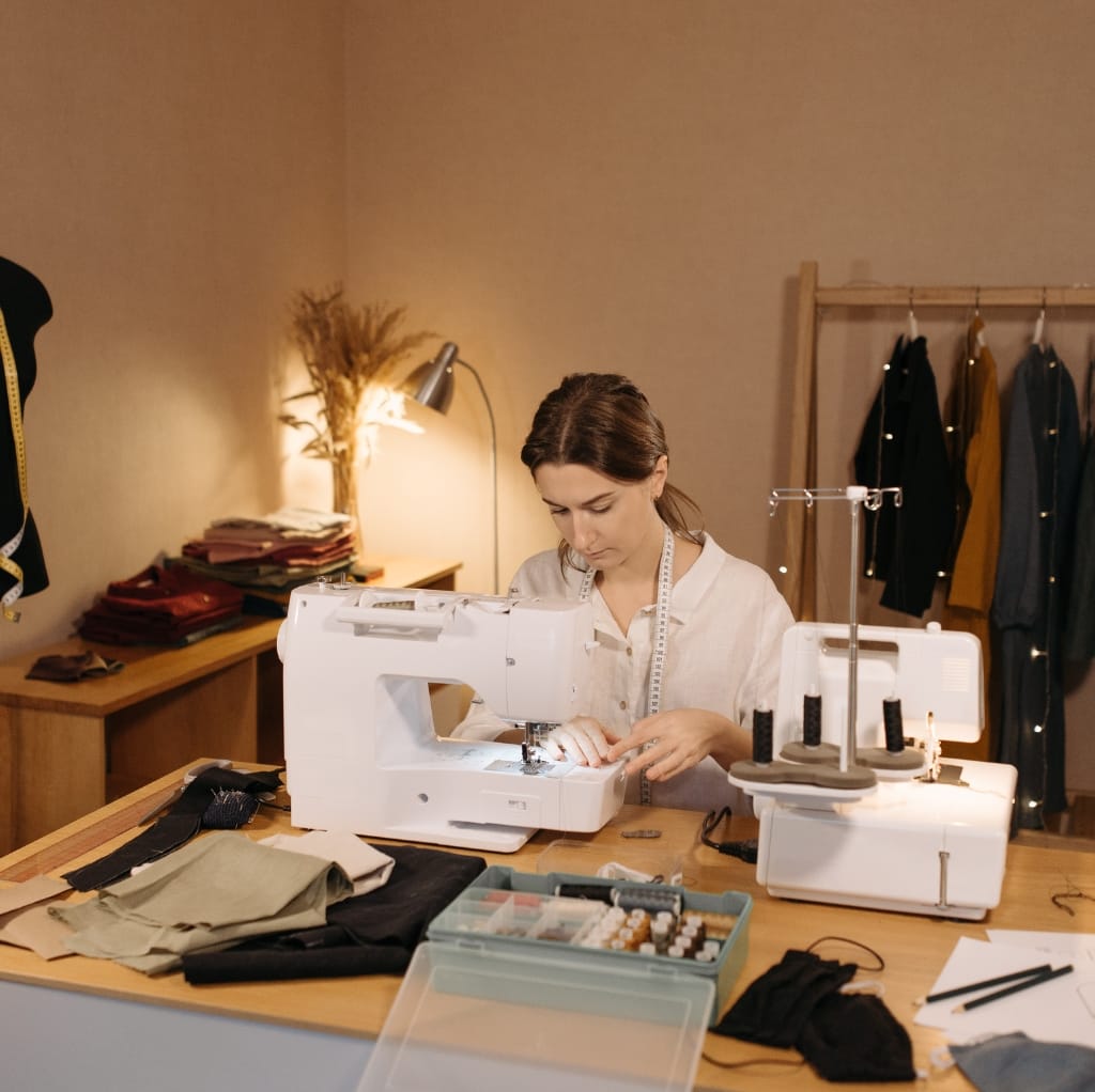 A woman is focused on sewing with a sewing machine in a well-lit workspace. The table has fabric, sewing tools, and thread organizers, with clothes hanging on a rack in the background.