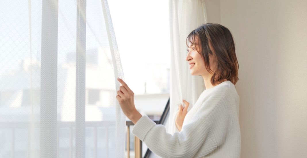 A young woman with shoulder-length brown hair, smiling, as she gently pulls back a sheer white curtain in a bright room.