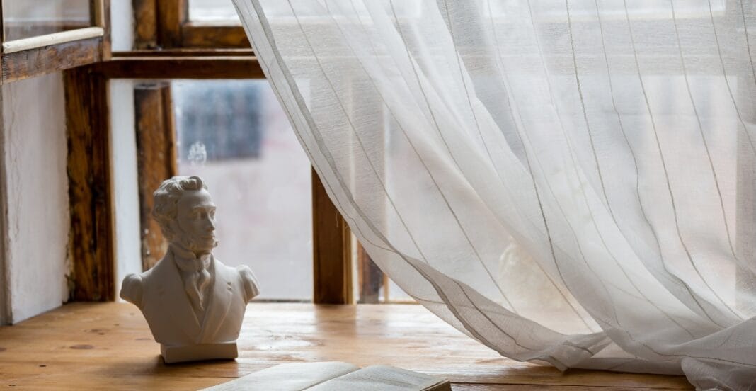 A white bust of a man with a beard and period clothing placed on a wooden surface next to a large, sheer curtain blowing in the breeze, with a window in the background.