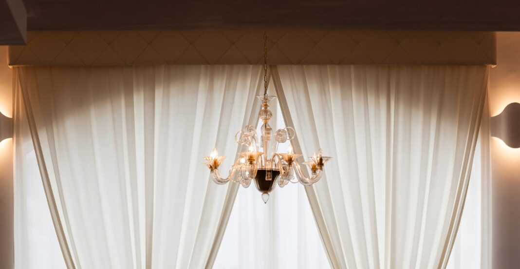 A chandelier hanging from the ceiling in front of a large window with white curtains.