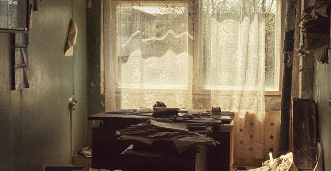 A cluttered, abandoned office with a dusty window and tattered curtains, a disorganized desk covered in papers, and peeling walls.