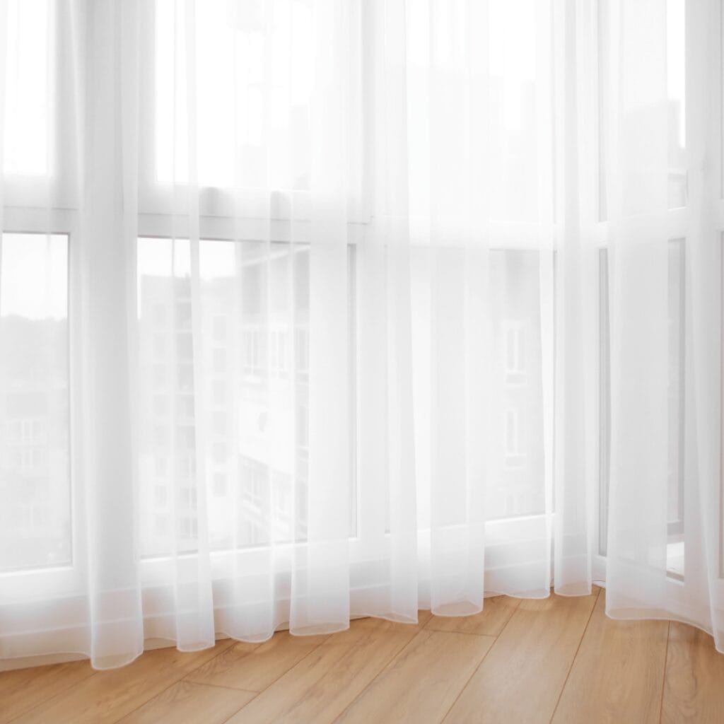 A bright, airy room with large windows covered by sheer white curtains, allowing diffused sunlight to enter. The floor is made of light-colored wood.