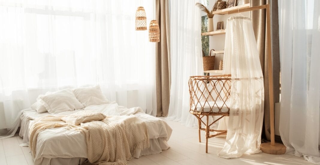 A bright, airy bedroom with white sheer curtains, a bed with cream-colored bedding and pillows, and a woven bassinet next to a wooden shelf with decorative items. The room has neutral tones and natural light.