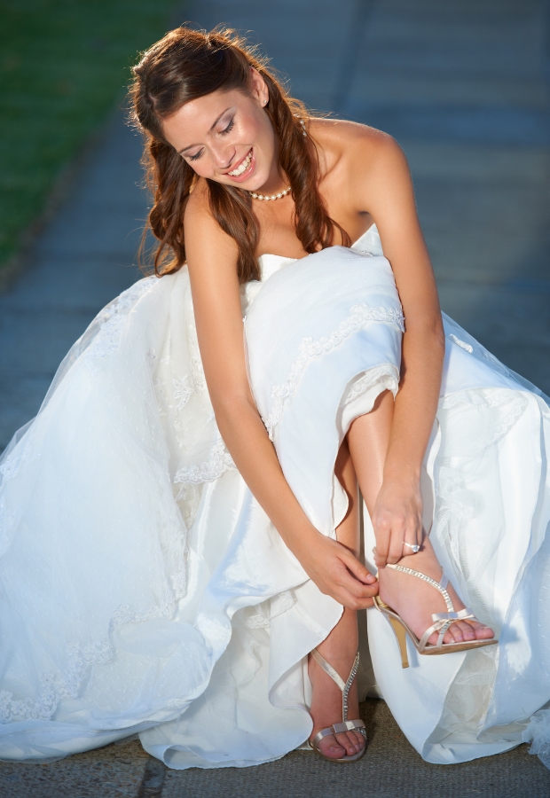A woman in a white wedding dress adjusts her high-heeled sandals, which feature delicate straps and a sparkling embellishment.