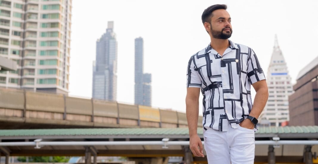 A man wearing a short sleeve button-down shirt with a geometric pattern, standing outdoors in an urban setting with tall buildings in the background.