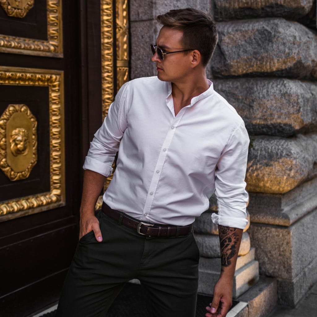 A man wearing a white short-sleeve button-down shirt, black pants, and sunglasses, standing outdoors near a building with ornate gold and dark wood details.