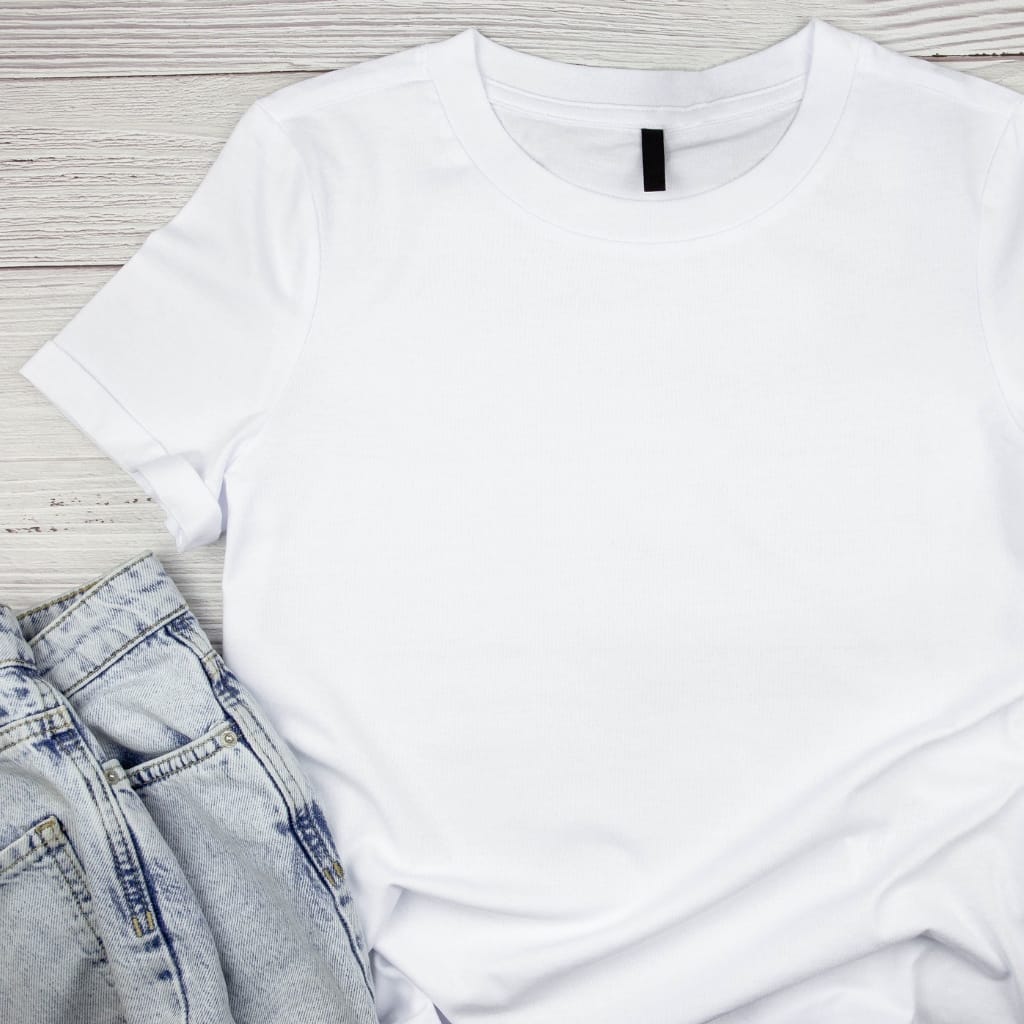 A plain white T-shirt laid flat on a light wooden surface, with a pair of light blue denim jeans partially visible at the bottom left.