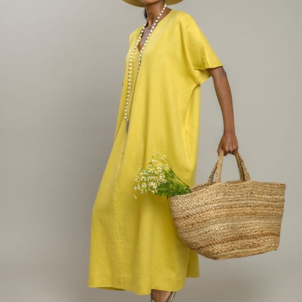 Woman wearing a yellow dress and a large straw hat, holding a woven basket with small white flowers, standing against a plain neutral background