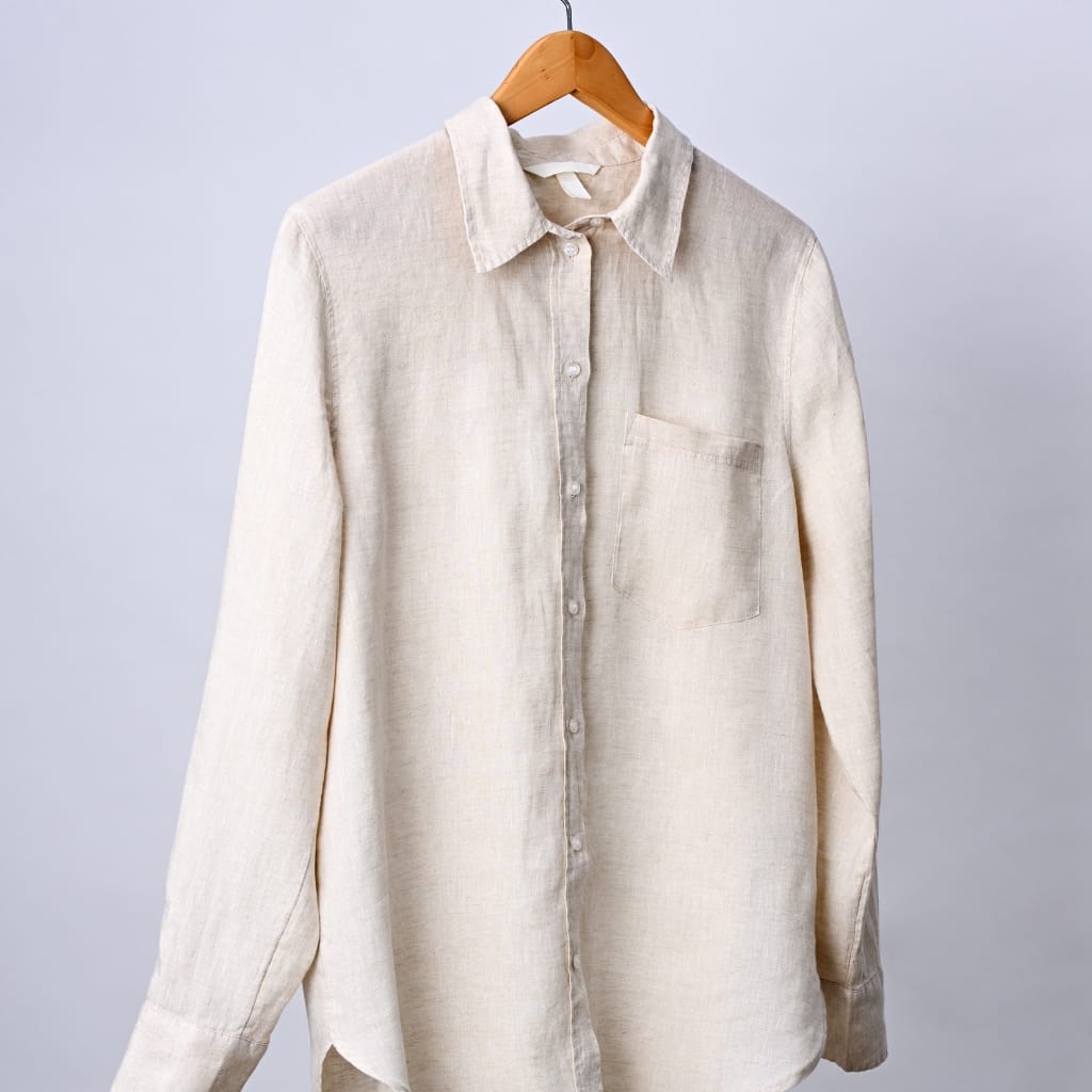 A light beige, long-sleeved button-up linen shirt hanging on a wooden hanger against a plain light background.