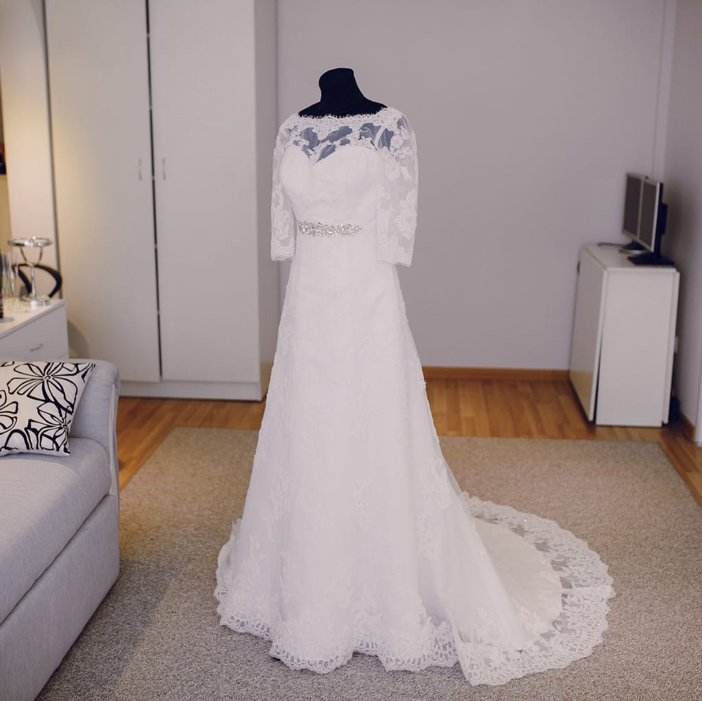 A white wedding dress displayed on a mannequin in a room with wooden flooring, a beige carpet, a gray sofa with a black and white patterned pillow, a white wardrobe, and a small TV on a white stand.
