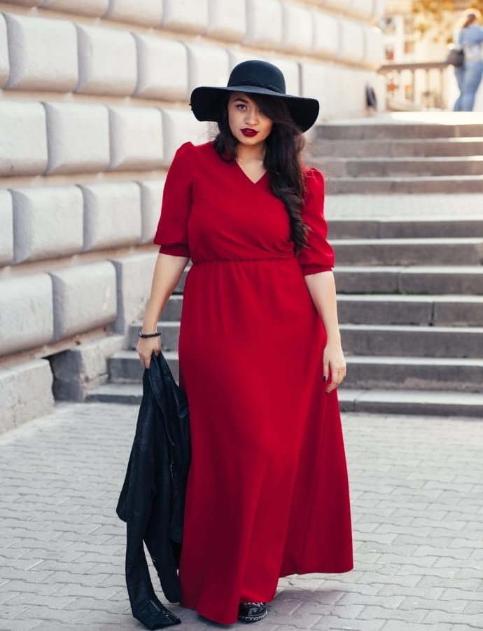 A woman dressed in a vibrant red maxi cotton dress, paired with a wide-brimmed black hat, holding a black jacket, walking outdoors near stone steps.