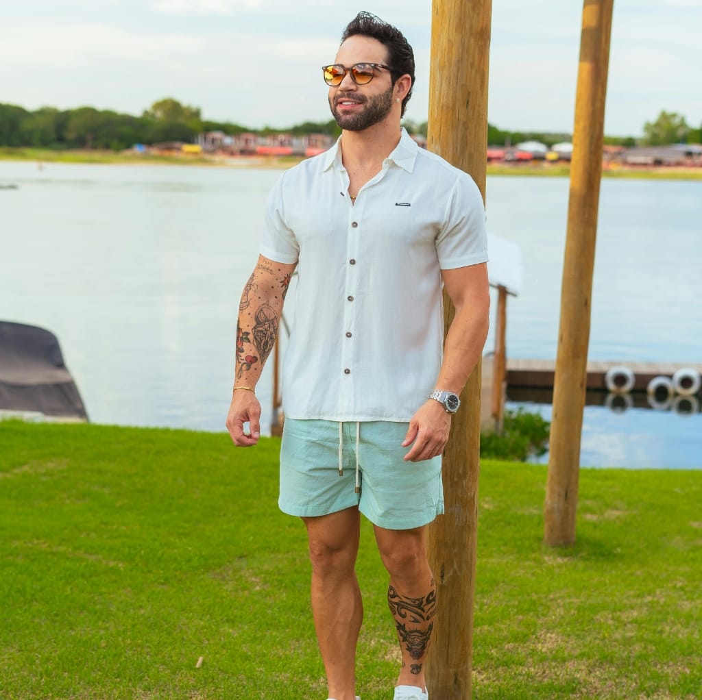 A man standing outdoors near a body of water, wearing a white linen short-sleeve shirt, light-colored shorts, sunglasses, and a wristwatch.