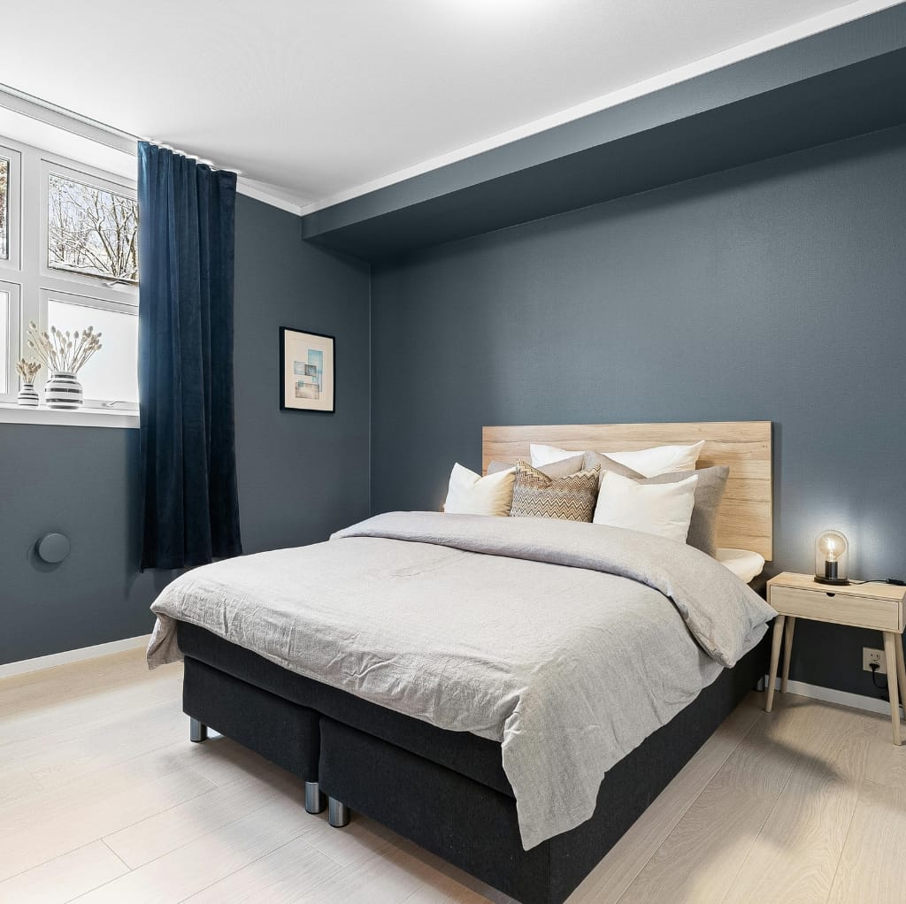 A modern bedroom featuring a neatly made bed with a beige duvet, multiple white and beige throw pillows, a light wood headboard, a minimal nightstand with a small lamp, and dark blue curtains next to a window with natural light. The walls are painted a muted blue, creating a cozy and stylish atmosphere.