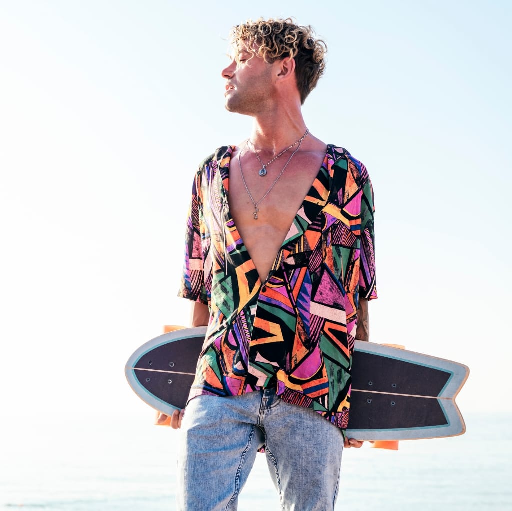 A stylish young man at the beach wearing a vibrant, colorful men's shirt open as a cover-up, holding a skateboard under his arm, with a bright sky and ocean in the background.