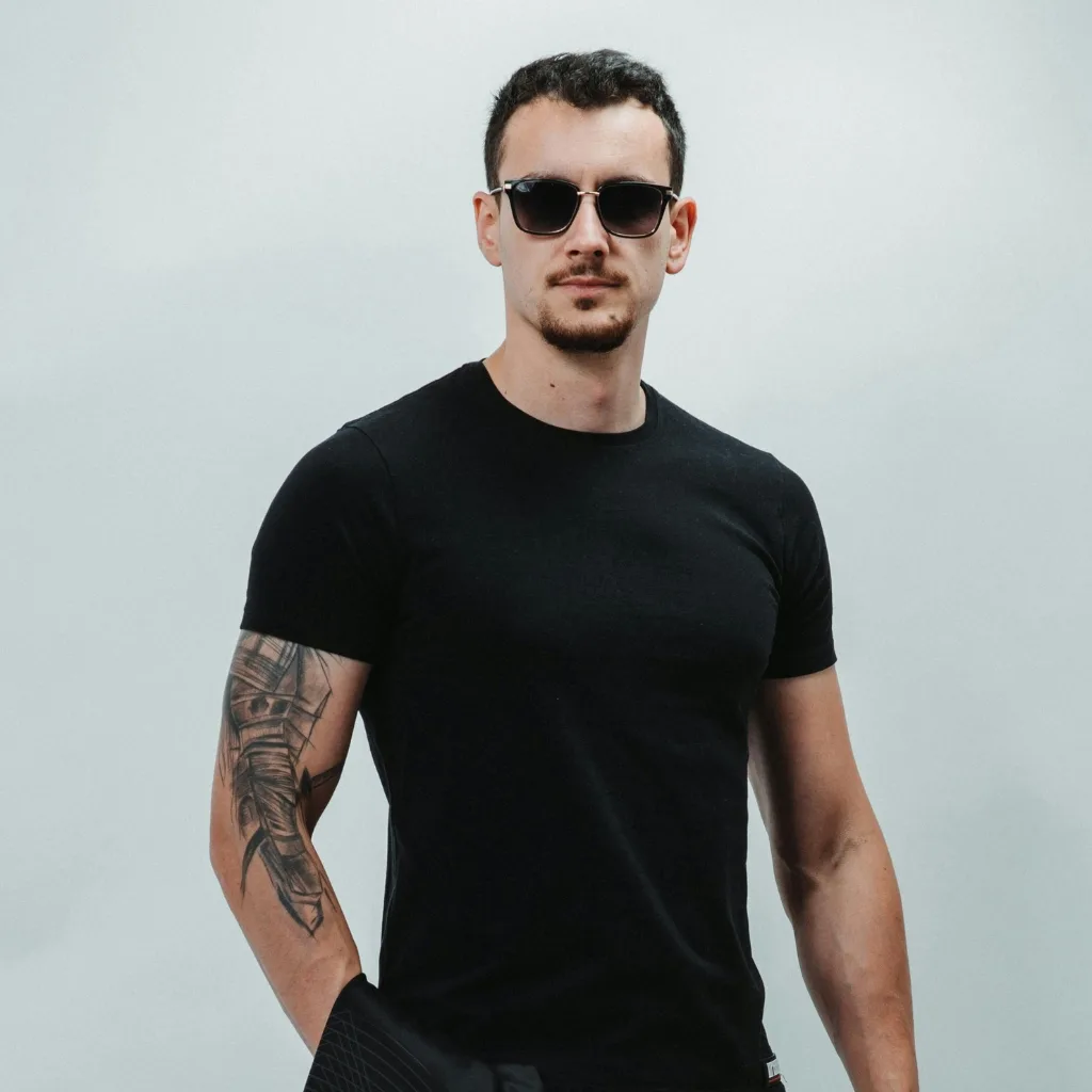 A man wearing sunglasses and a black T-shirt with tattooed arm, standing against a light gray background.