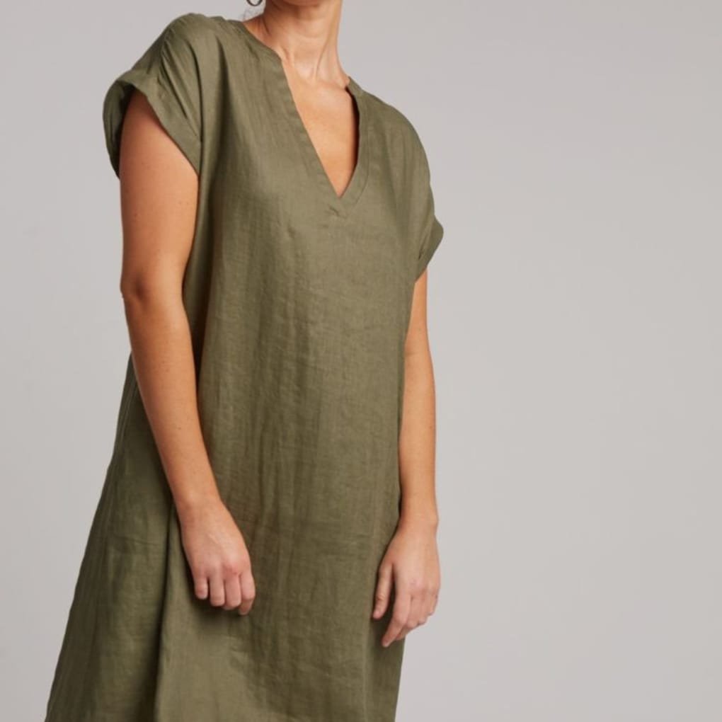 Woman wearing an olive-green, loose-fitting, short-sleeved linen dress with a V-neckline, standing against a plain neutral background.