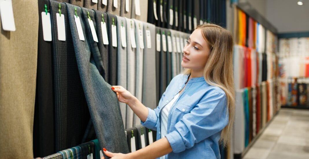 A young woman in a blue shirt shopping for fabric or textiles at a store, holding a swatch and examining it against other fabric rolls organized on a display.