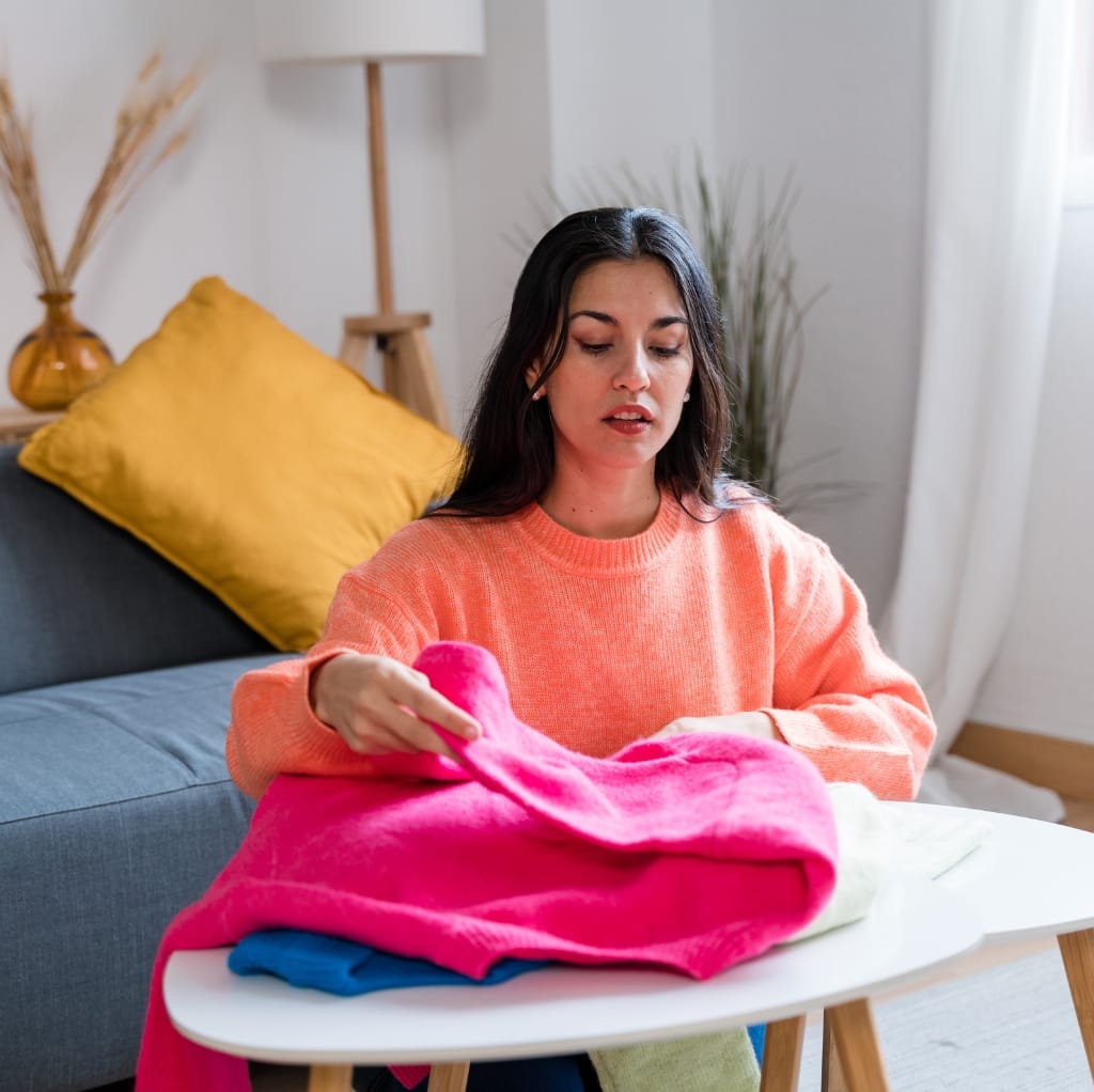 A young woman with dark hair, wearing an orange sweater, is sitting at a small table sorting through a pile of colorful fleece fabrics in a bright, cozy room.