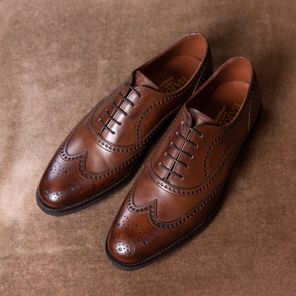 A pair of polished tan brogue or derby shoes with decorative perforations and stitching, placed on a brown textured surface.