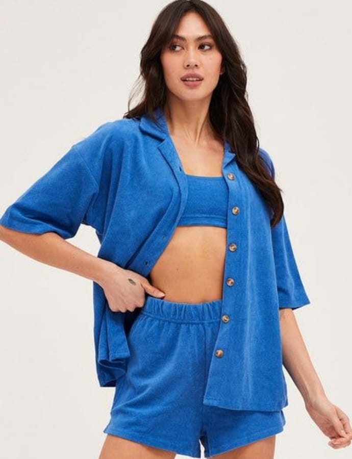 A woman wearing a matching blue terrycloth short-sleeve shirt and shorts, with an unbuttoned shirt revealing a blue sports bra beneath, standing against a plain background.