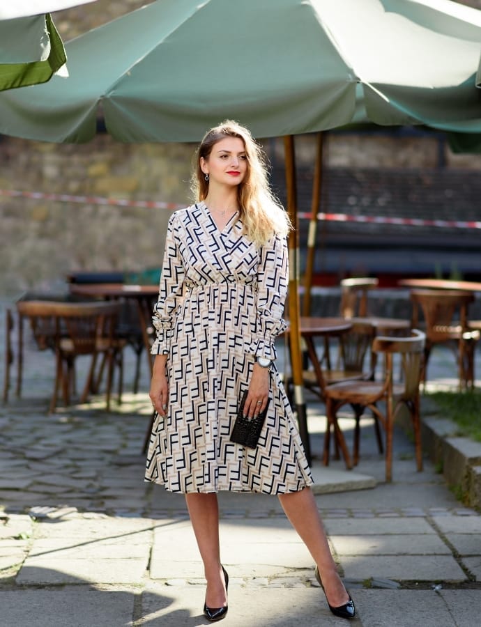 A stylish woman standing outdoors under a large green umbrella, wearing a patterned midi cotton dress with long sleeves, black high heels, and holding a small black clutch. The background features outdoor seating and stone pavement.