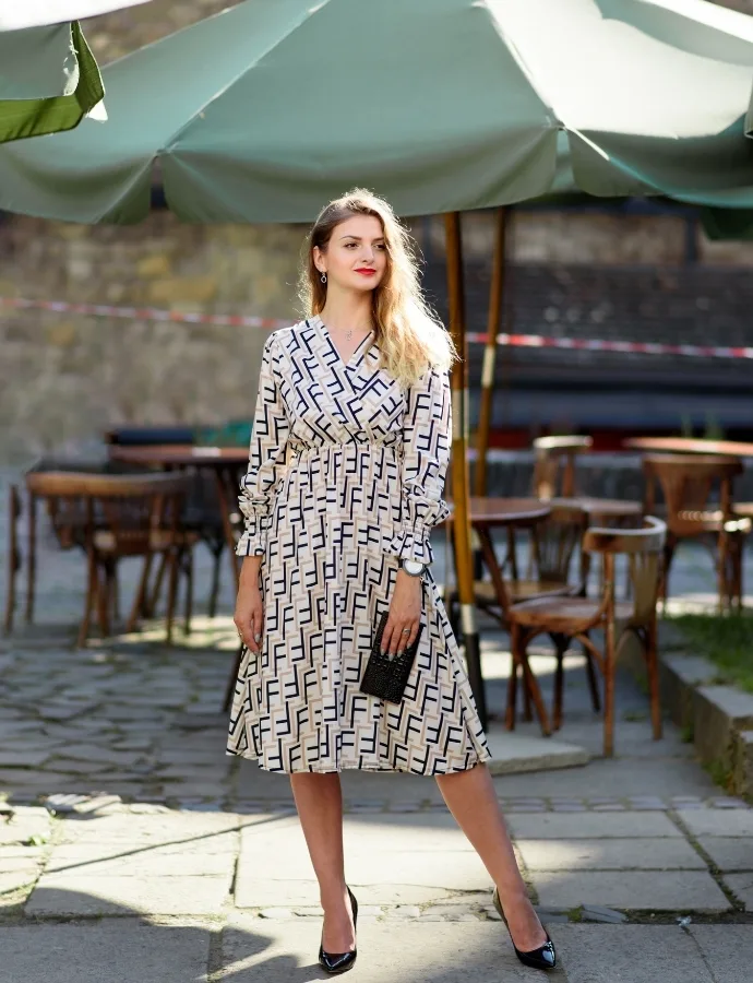 A stylish woman standing outdoors under a large green umbrella, wearing a patterned midi cotton dress with long sleeves, black high heels, and holding a small black clutch. The background features outdoor seating and stone pavement.