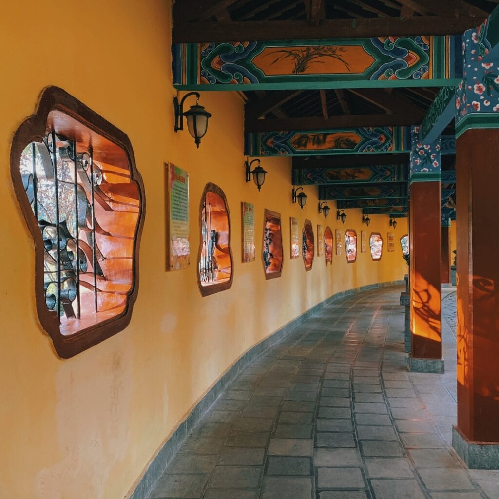 A curved corridor with yellow walls, decorated with framed art and ornamental windows with wrought iron grilles. The ceiling has colorful, traditional patterns, and there are black lantern-style wall sconces along the wall. The floor is tiled with a mix of rectangular and square tiles.