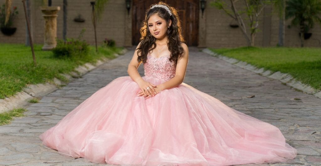 A young girl wearing a voluminous pink tulle prom dress sitting on a stone pathway in an outdoor garden setting.