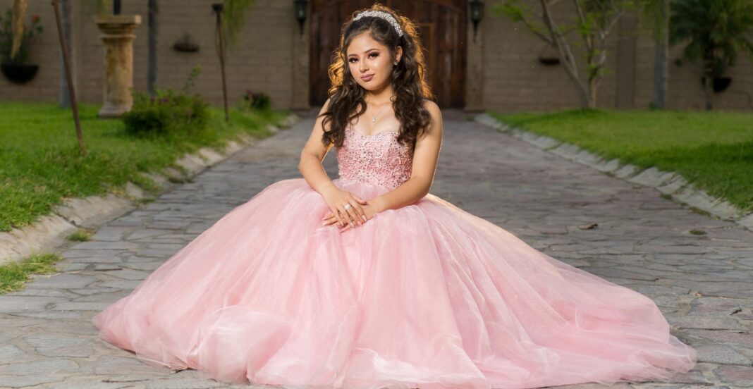 A young girl wearing a voluminous pink tulle prom dress sitting on a stone pathway in an outdoor garden setting.