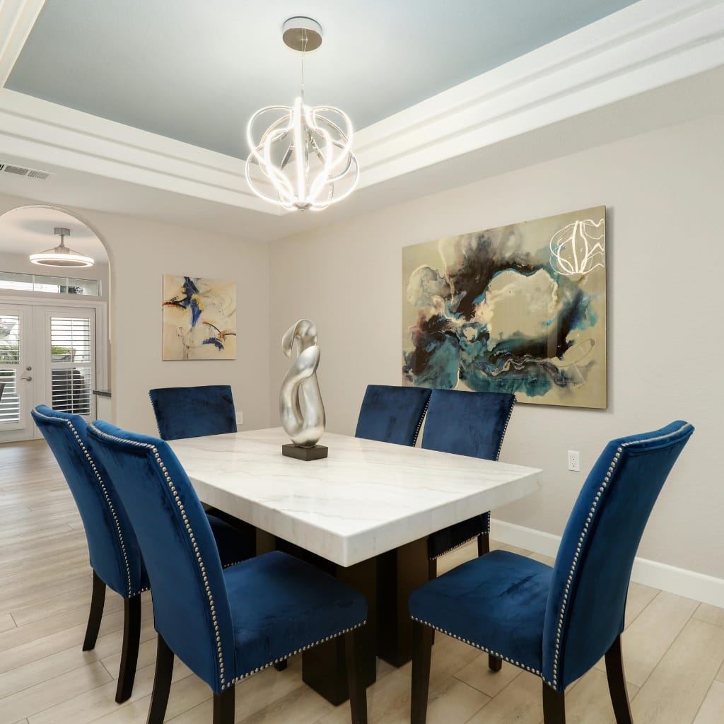 A modern dining room with a white marble-top table surrounded by six blue velvet upholstered chairs, complemented by contemporary artwork and a stylish light fixture.