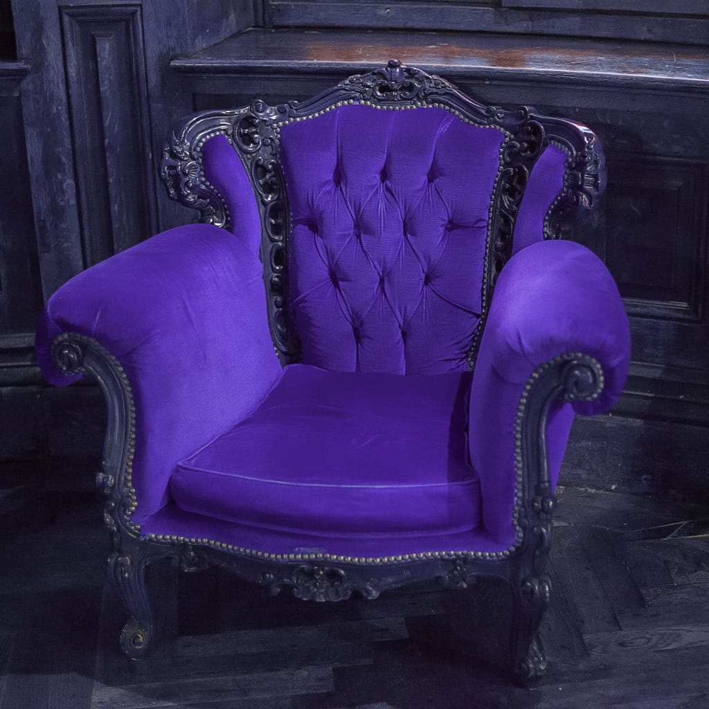 A vintage-style armchair with ornate wooden carvings and deep purple velvet upholstery, set against a dark wooden wall.