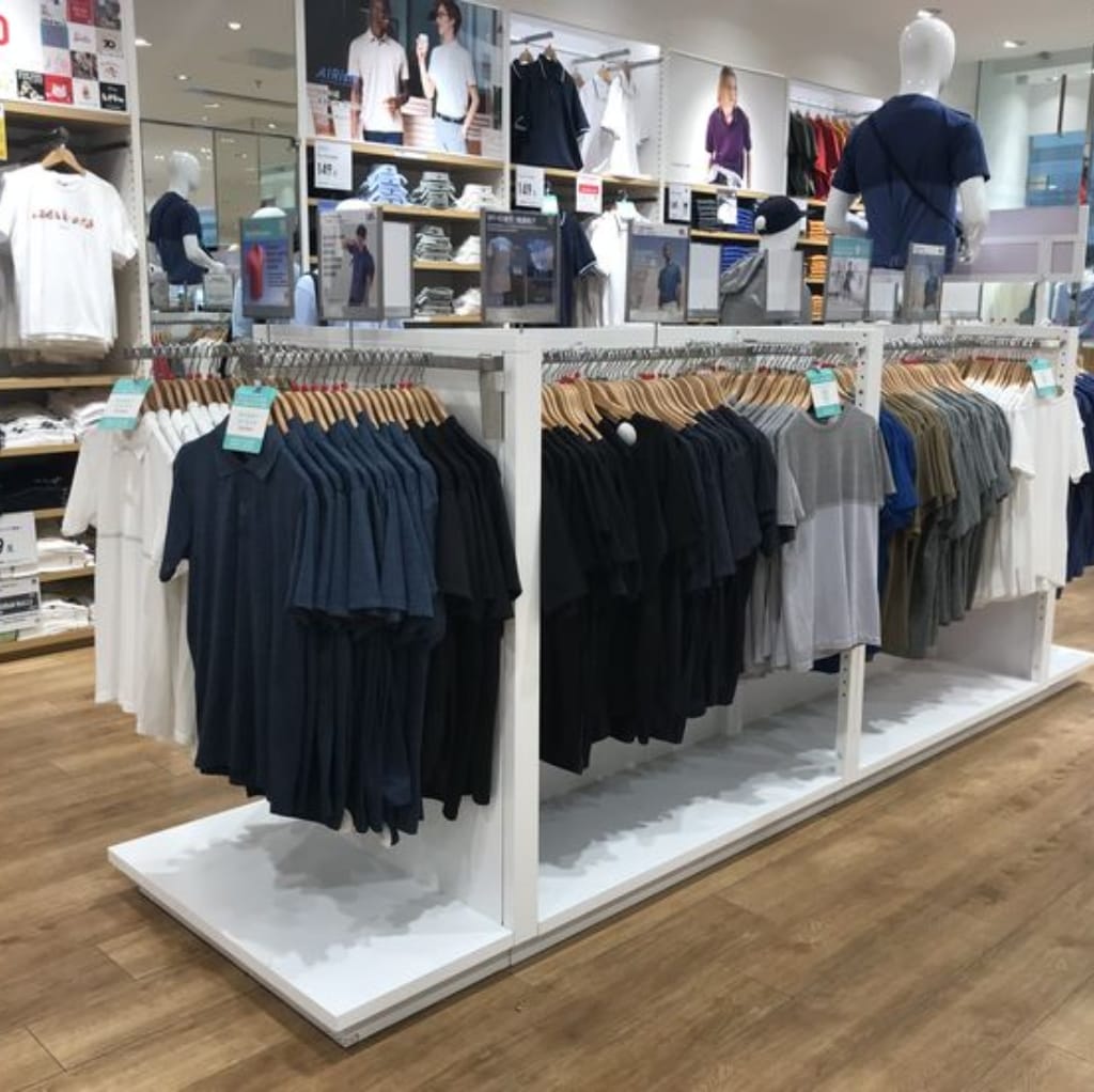 Clothing display in a Uniqlo store featuring racks of polo shirts in various colors including navy blue, black, gray, white, and beige, with shelves of folded shirts and mannequins dressed in casual apparel in the background.